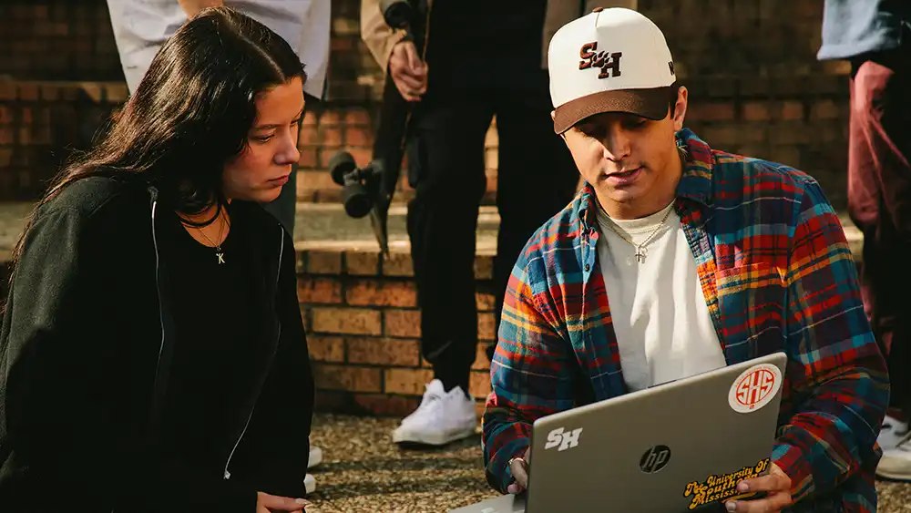 Two college students sit outside on a sunny day, looking at a laptop together. The student on the right wears a Sam Houston State University cap, colorful plaid shirt, and jeans, and is showing something on the screen to the other student. The student on the left wears all black and listens attentively. The laptop is decorated with SHSU-related stickers. Brick steps and other students with skateboards are visible in the background.