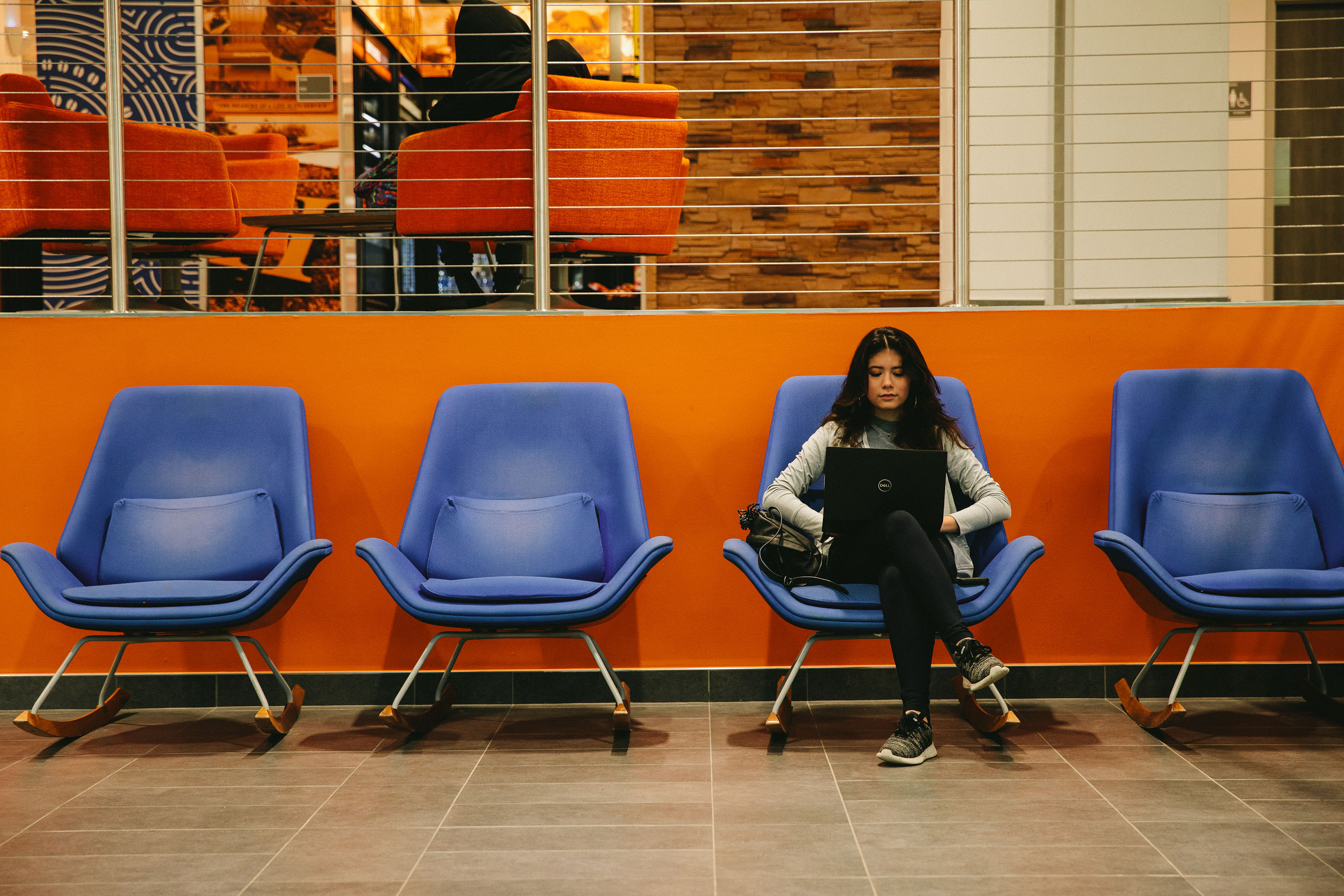 A person sitting in a blue chair in a waiting area with an orange wall, using a laptop. The area includes three other identical blue chairs. Behind the orange wall is a glass partition revealing another room with orange chairs and wooden wall paneling.
