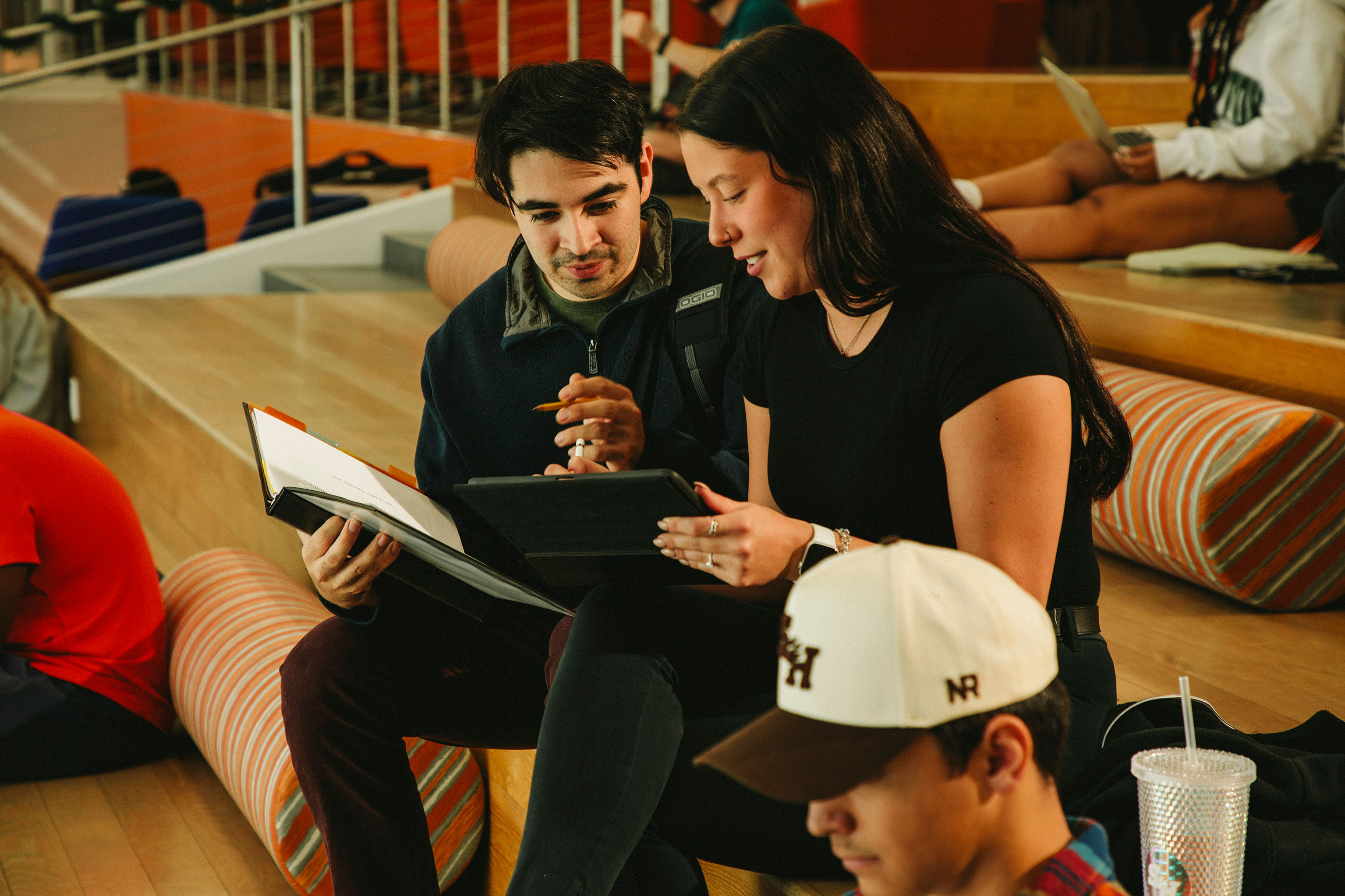 ​​Two students sit on tiered seating indoors, collaborating over a tablet and on an open binder. The student on the left gestures with a pencil while the student on the right holds the tablet. A third student in the foreground wears a SHSU cap and focuses on his own work. Other students are seated in the background.​ 