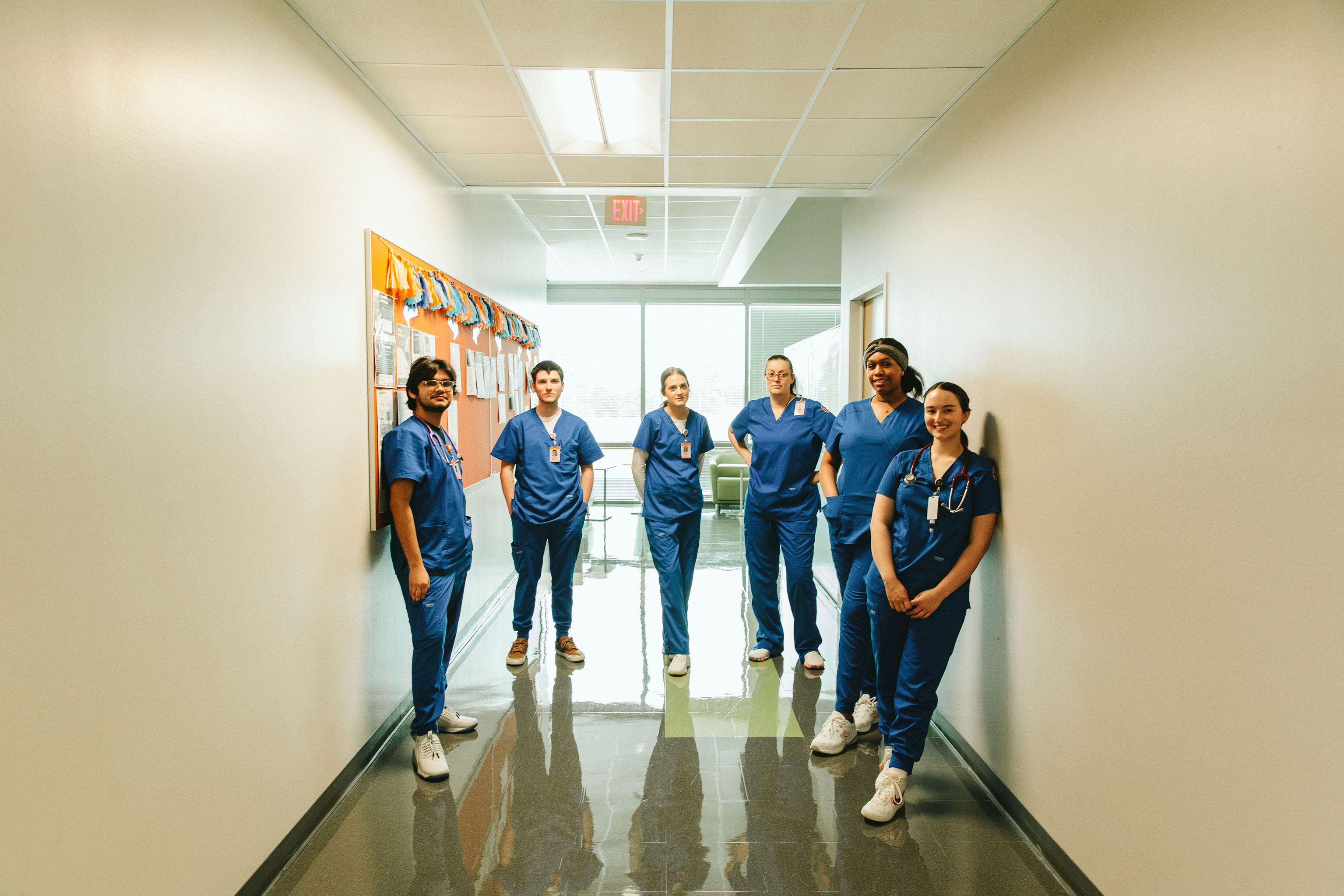 Six individuals in blue medical scrubs and white shoes stand in a semi-circle in a bright hallway, facing the camera. The hallway features light-colored walls, a reflective floor, and a decorated bulletin board on the left. An exit sign and large windows in the background allow natural light into the space.