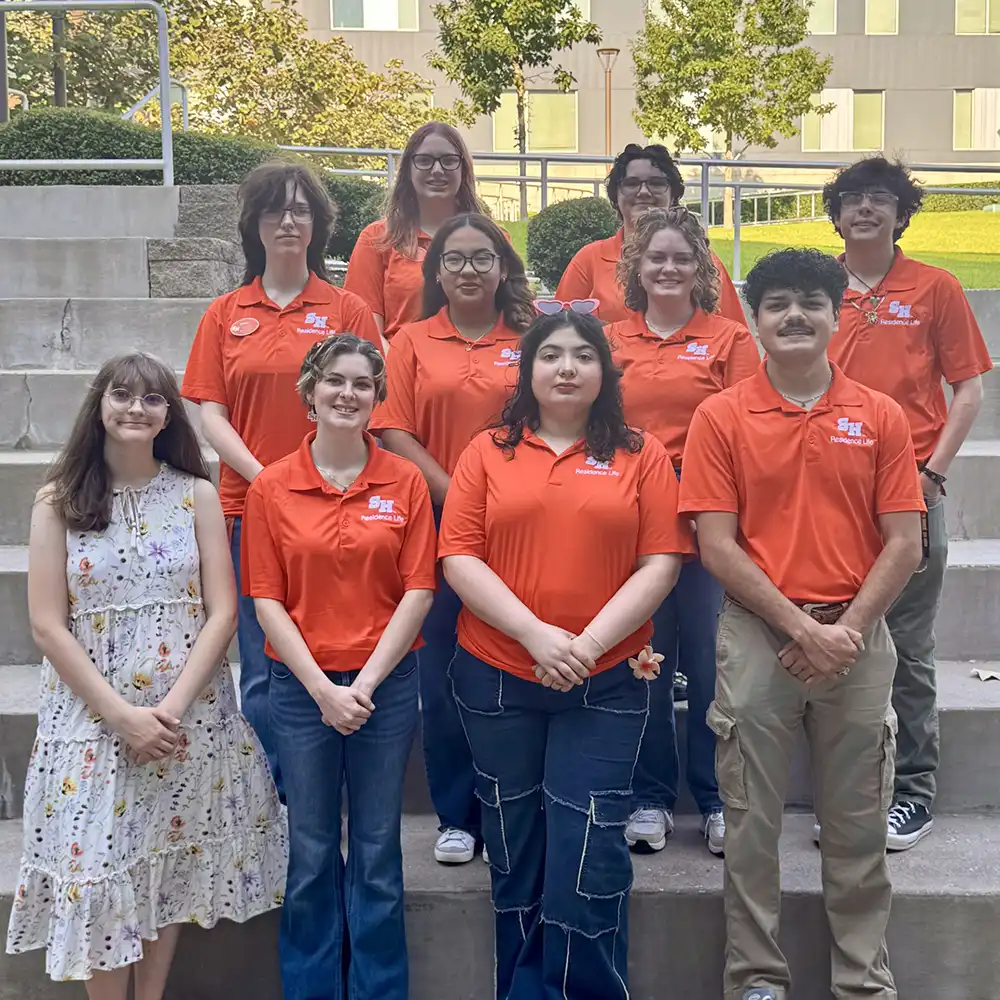 Group photo of Sam Houston State University Residence Life hall staff standing on outdoor steps in front of a residence hall. Most of the staff are wearing matching orange SHSU Residence Life shirts, while one person in the front row is wearing a white floral dress. The group is smiling and posing together for the photo.
