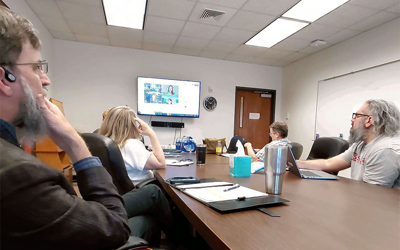 Faculty and staff gathered around a conference table discussing instructional design with other faculty and staff joining in over zoom on a television in the background.