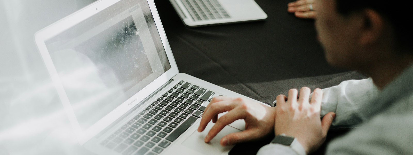 An over-the-shoulder view of a person using a silver laptop on a black tablecloth. The person's left hand is on the trackpad. Another laptop and a person's hands are visible in the blurred background, suggesting a meeting or workspace environment.