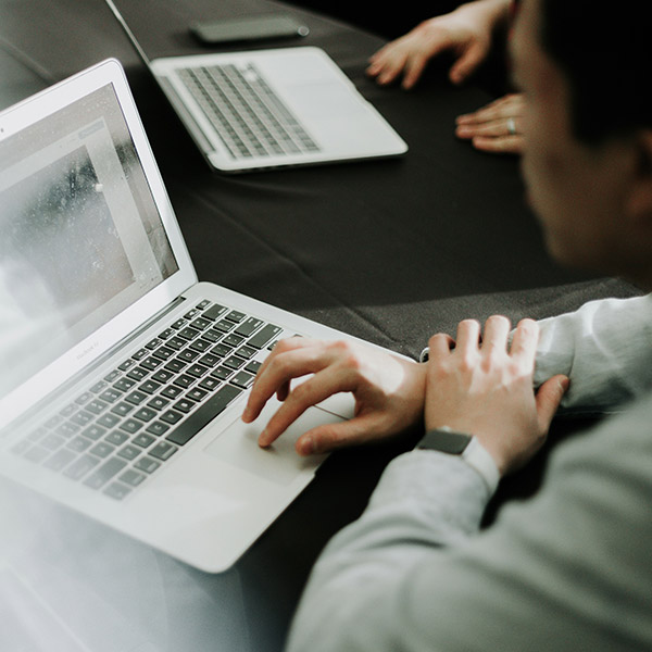 An over-the-shoulder view of a person using a silver laptop on a black tablecloth. The person's left hand is on the trackpad. Another laptop and a person's hands are visible in the blurred background, suggesting a meeting or workspace environment.