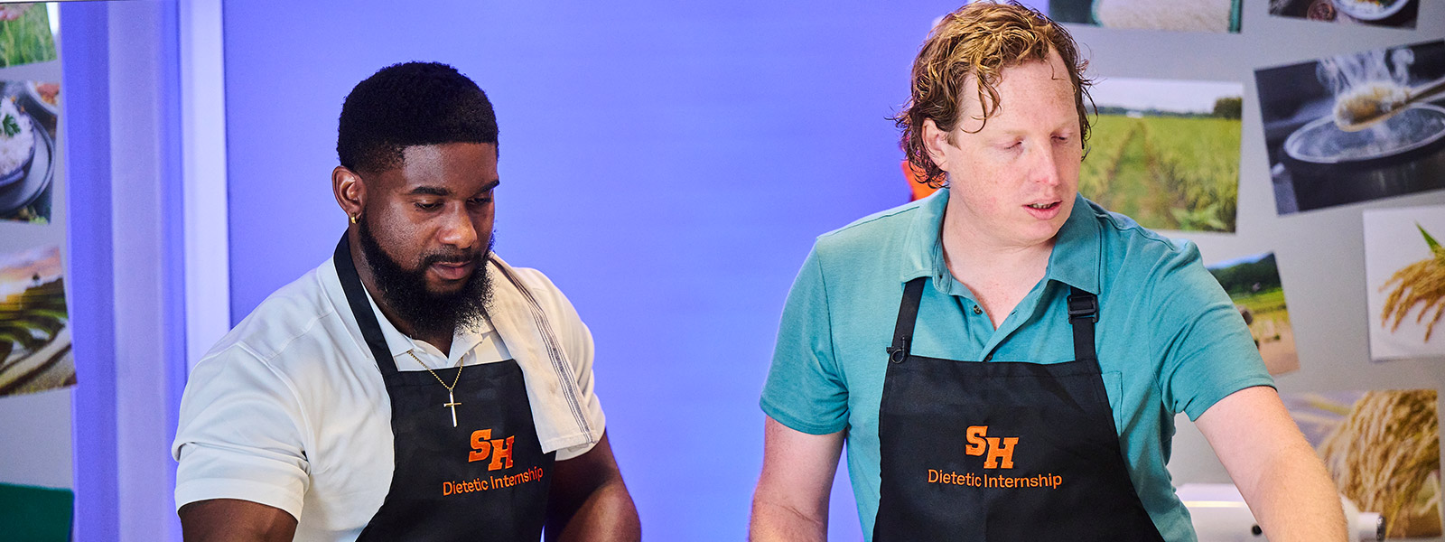 Two men wearing black aprons with the "SHSU Dietetic Internship" logo working in a professional kitchen. The man on the left is holding a metal mixing bowl, and the man on the right is looking toward a workspace with fresh vegetables, including green bell peppers and carrots.