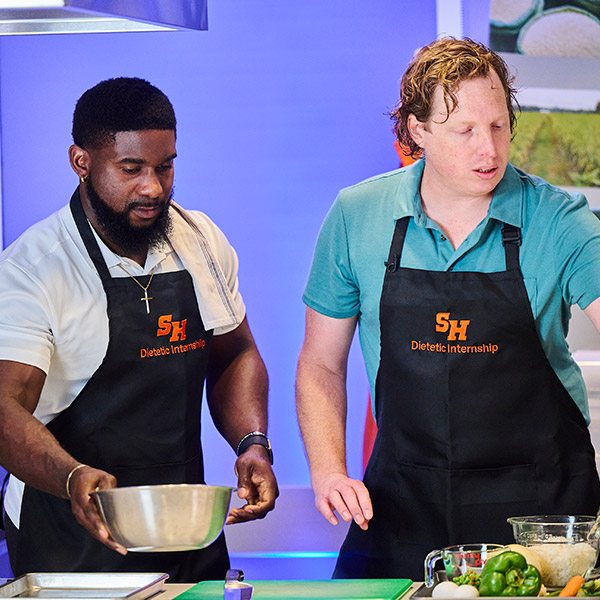 Two men wearing black aprons with the "SH Dietetic Internship" logo working in a professional kitchen. The man on the left is holding a metal mixing bowl, and the man on the right is looking toward a workspace with fresh vegetables, including green bell peppers and carrots.