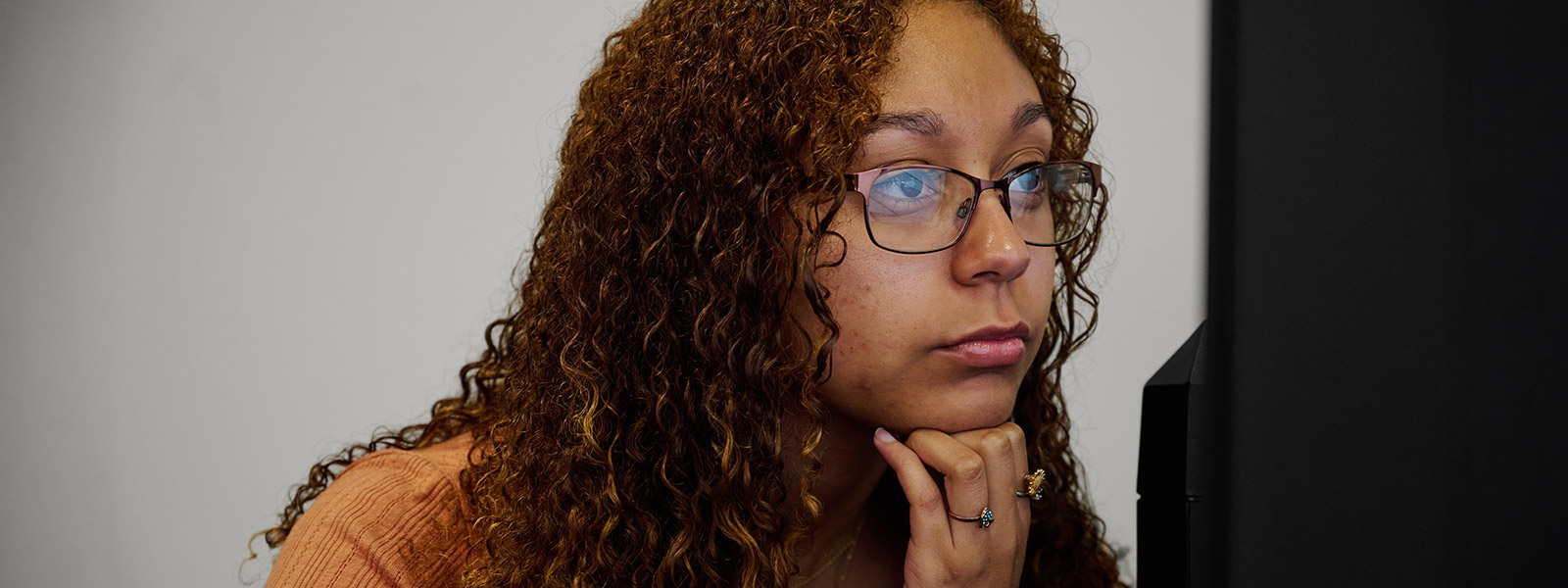 A close-up profile shot of a young woman with long, curly brown hair and glasses. She is resting her chin on her hand with a thoughtful expression while looking at a computer screen that is partially visible on the right side of the frame.