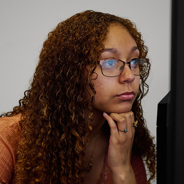 A close-up profile shot of a young woman with long, curly brown hair and glasses. She is resting her chin on her hand with a thoughtful expression while looking at a computer screen that is partially visible on the right side of the frame.