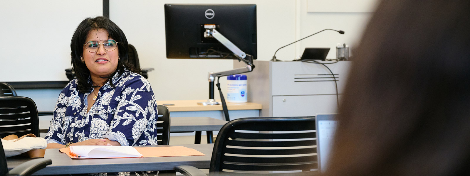 A female professor with dark hair and glasses, wearing a blue and white floral patterned blouse, sits at a desk in a classroom. She is looking toward someone off-camera with an attentive and pleasant expression. On the desk in front of her is a notepad and a folder. The background features student desks, black office chairs, and a computer monitor mounted on an adjustable arm.