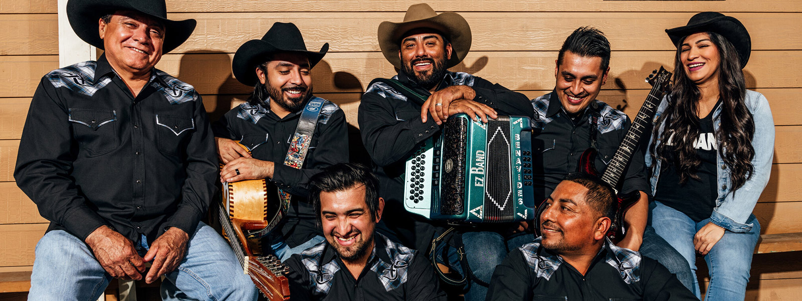 A group portrait of seven members of a band. They are dressed in matching black western shirts with decorative silver patterns on the shoulders, jeans, and cowboy hats. Several members hold instruments, including an accordion and a snare drum. They are smiling and laughing together against a tan wooden wall decorated with two metal fish skeleton art pieces.