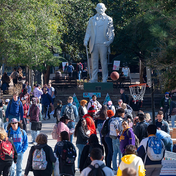 A crowded outdoor campus plaza at Sam Houston State University. Students are walking in various directions. In the center stands a large white statue of Sam Houston. To the right, a basketball is captured mid-air near a hoop. Trees and campus buildings are visible in the background under bright sunlight.