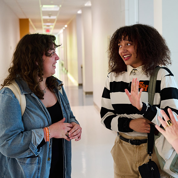 Two female students engaged in a friendly conversation in a brightly lit, white-walled hallway. The student on the left has long brown hair and wears a denim jacket. The student on the right has curly dark hair and is wearing a black-and-white striped shirt and khaki pants, gesturing with her hand while she speaks.