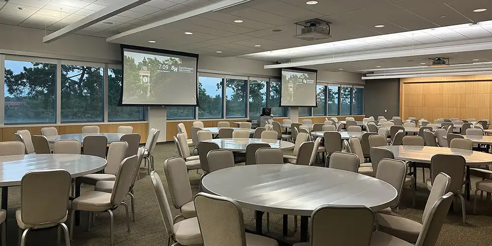 Sam Houston Room with a setup of round tables and with the projector screens down.